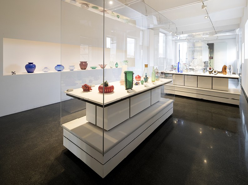 A modern gallery interior with white walls and dark floors, displaying colorful Murano glass art pieces in clear vitrines and on pedestals.