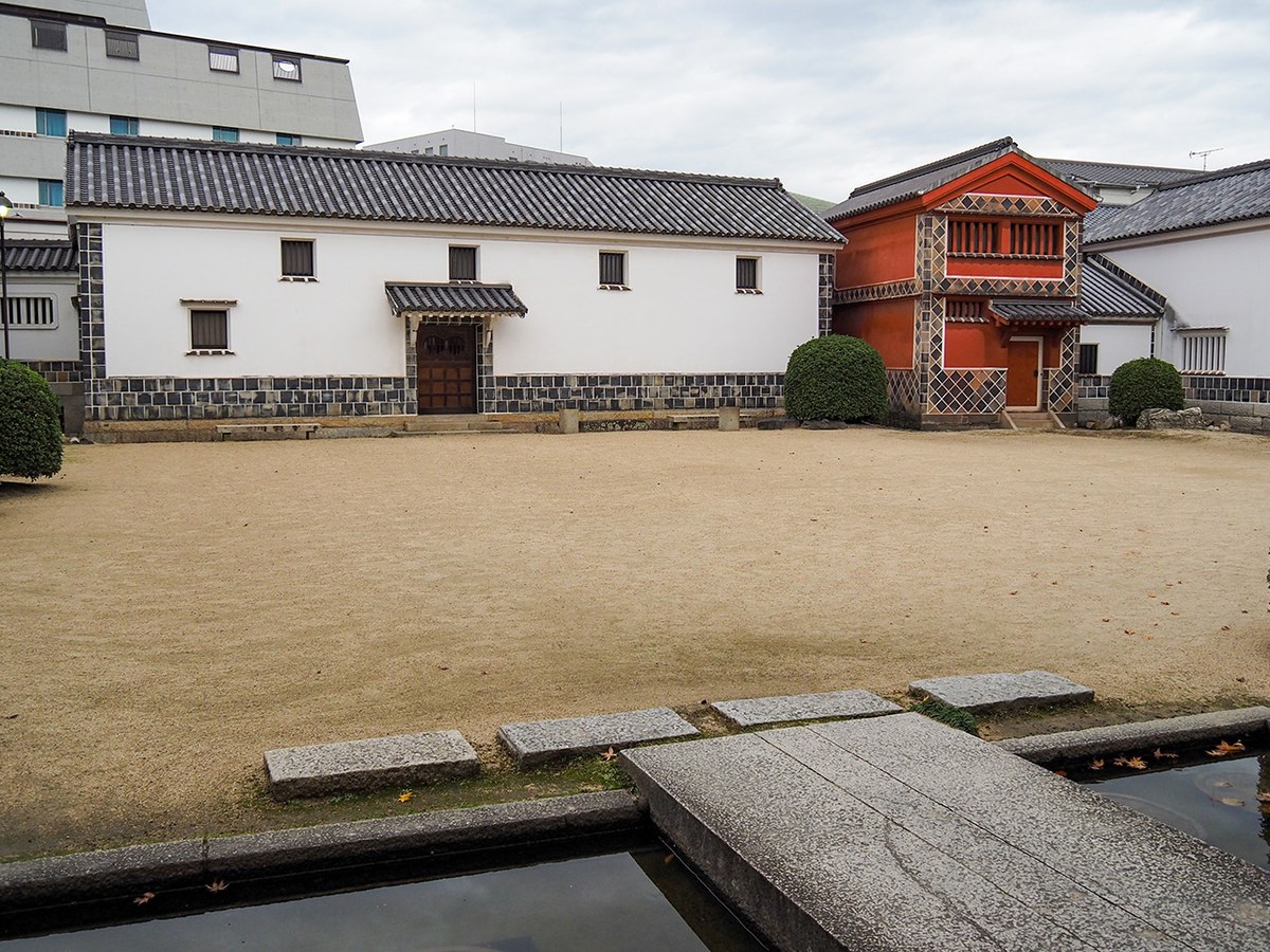 Exterior courtyard of traditional Japanese buildings with white walls, dark tiled roofs, and a distinctive red-painted structure, surrounded by