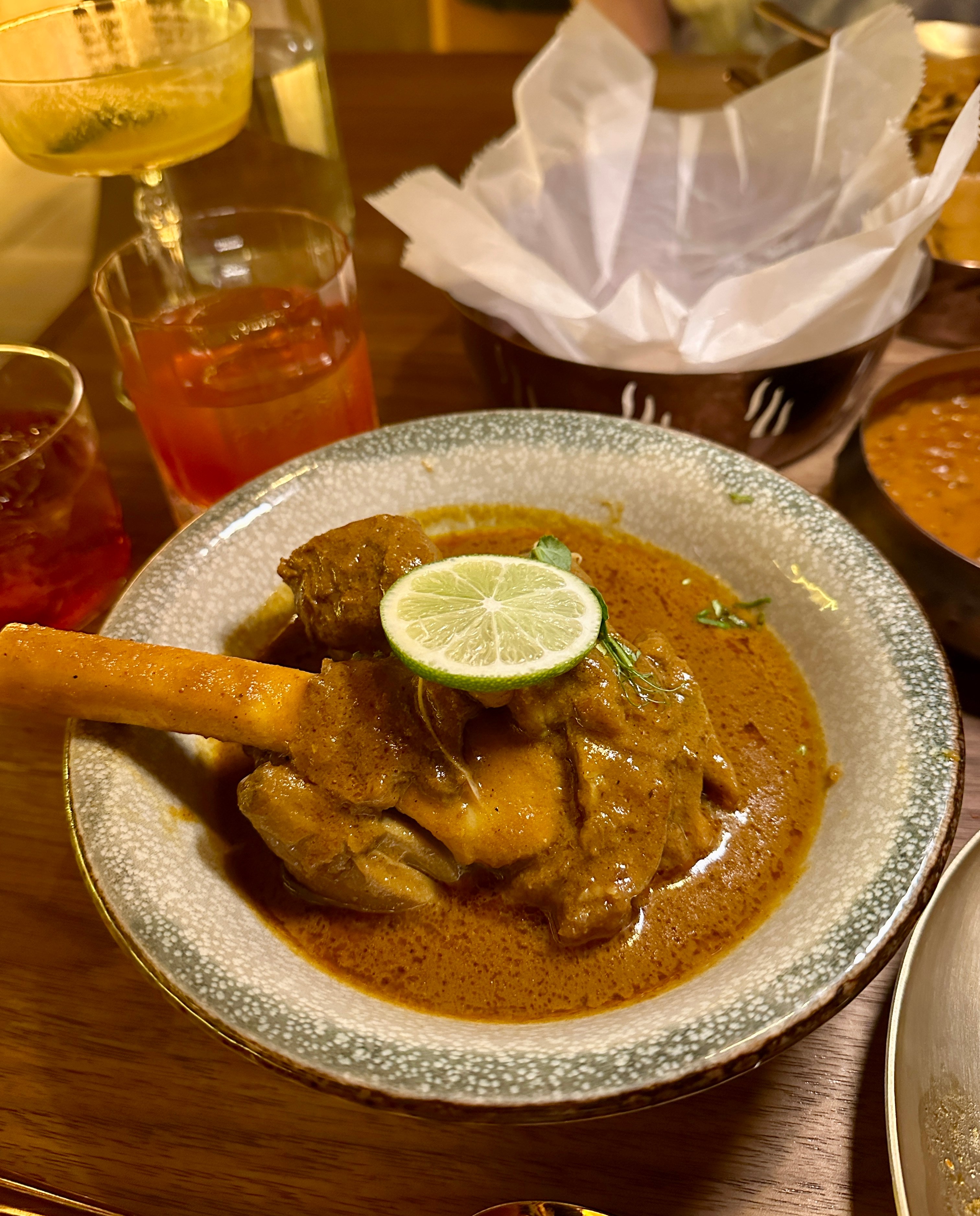 A speckled gray bowl containing a golden-brown curry with meat pieces, topped with a lime slice and fresh herbs, with a wooden spoon resting in the dish.