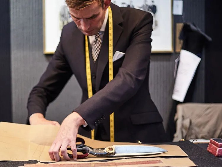 A tailor in a dark suit with a yellow measuring tape around his neck cuts a paper pattern on a workbench using large shears.