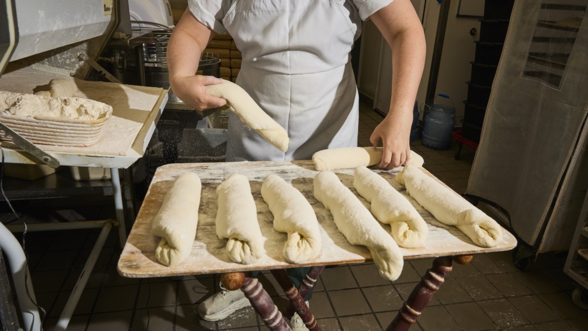 A baker in white chef's uniform holding shaped bread dough in a professional bakery kitchen