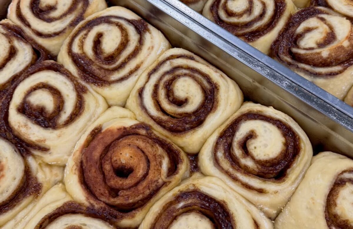 Close-up of multiple cinnamon scrolls on baking trays showing hand-rolled spiral layers with dark cinnamon filling