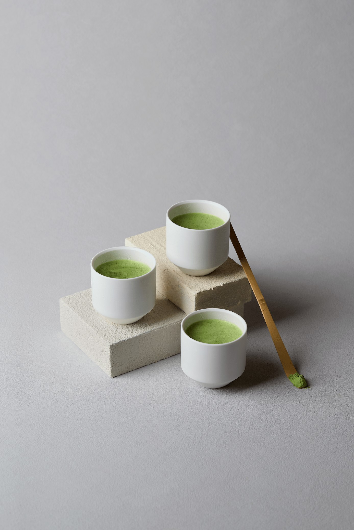 Three white cups containing bright green matcha tea arranged on cream-colored stone blocks with a wooden bamboo whisk beside them.