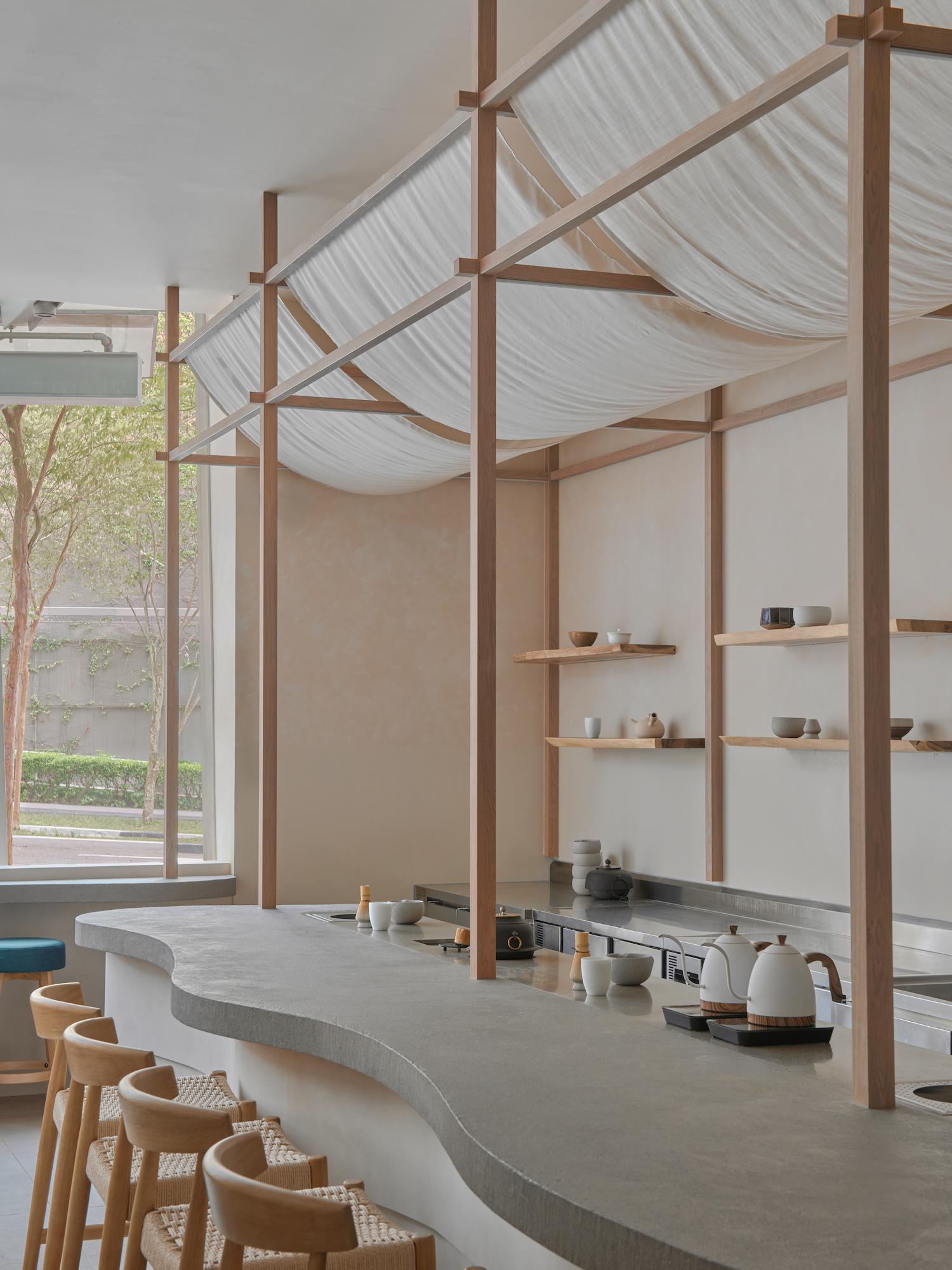 A minimalist matcha cafe interior with curved concrete counter, wooden structural frame with draped fabric canopy, and shelving displaying tea wares; 