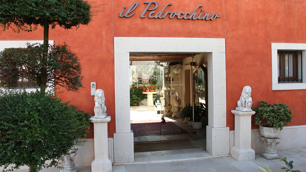The red-stucco facade of Ristorante Il Pedrocchino, featuring a stone-framed glass entrance flanked by lion statues and lush greenery.