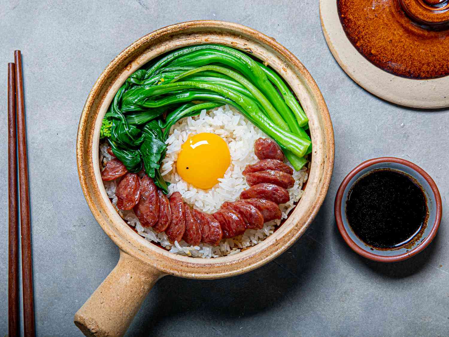 Clay pot with rice, sausage, greens, egg, and soy sauce on a gray surface.