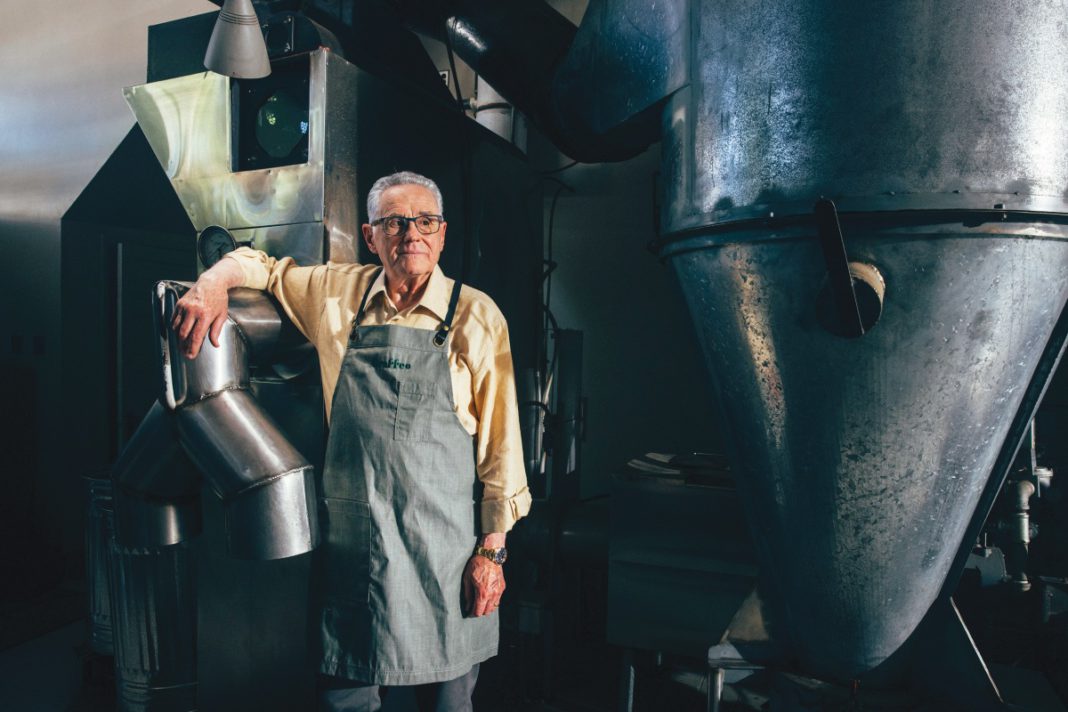 An older man wearing glasses, a yellow shirt, and gray apron stands in an industrial coffee roasting facility with large metal roasting equipment; the