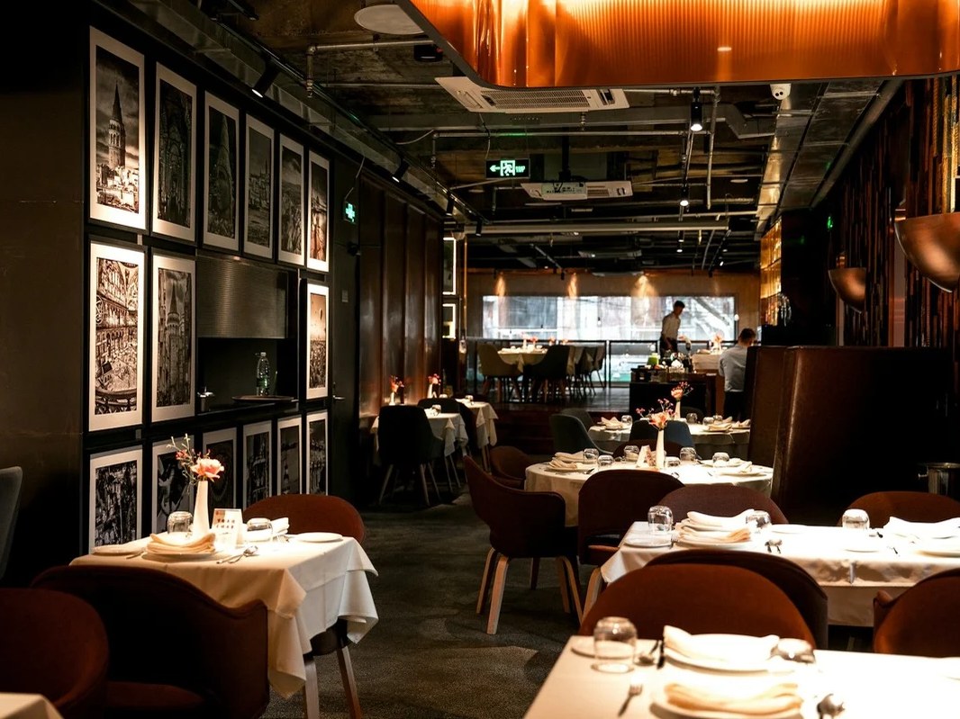 An upscale restaurant dining room with warm lighting, dark walls featuring framed black-and-white photographs, white-tablecloth tables with brown