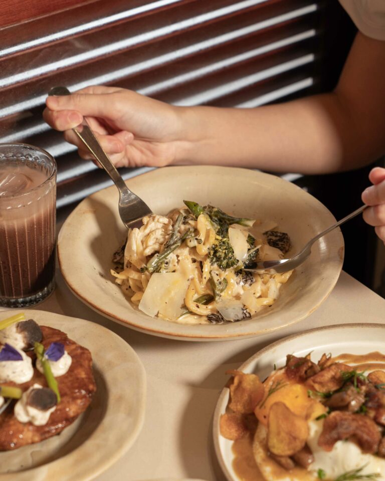 A person's hand holds a fork over a bowl of pasta with leafy greens, with other plates of food and a glass of beverage visible on the table.