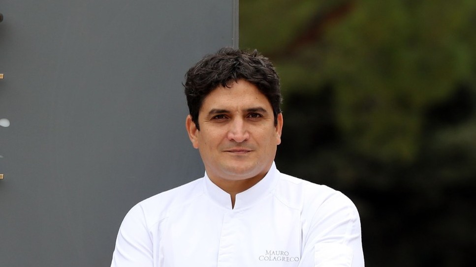 A man in a white chef's jacket with 'Mauro Colagreco' embroidered on the chest stands with arms crossed in front of a large sign reading 'Mirazur / Re