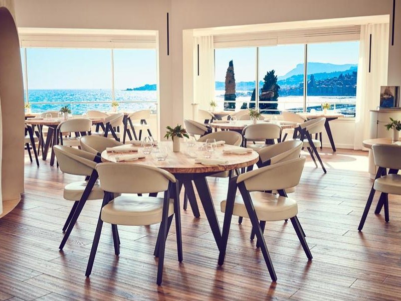 A bright, modern restaurant dining room with round wooden tables, cream upholstered chairs, and floor-to-ceiling sea-facing windows revealing a Medite