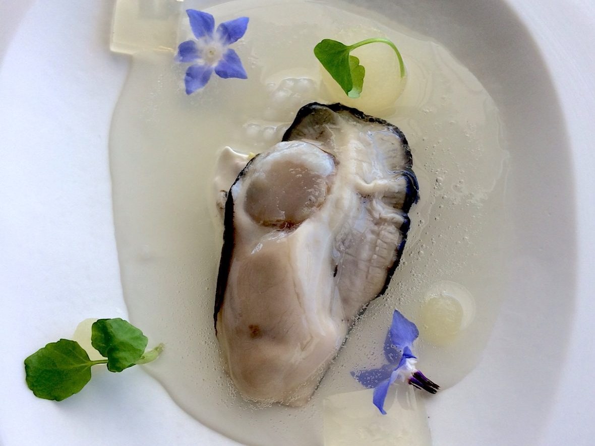 Oyster on a plate with edible flowers and a clear liquid.