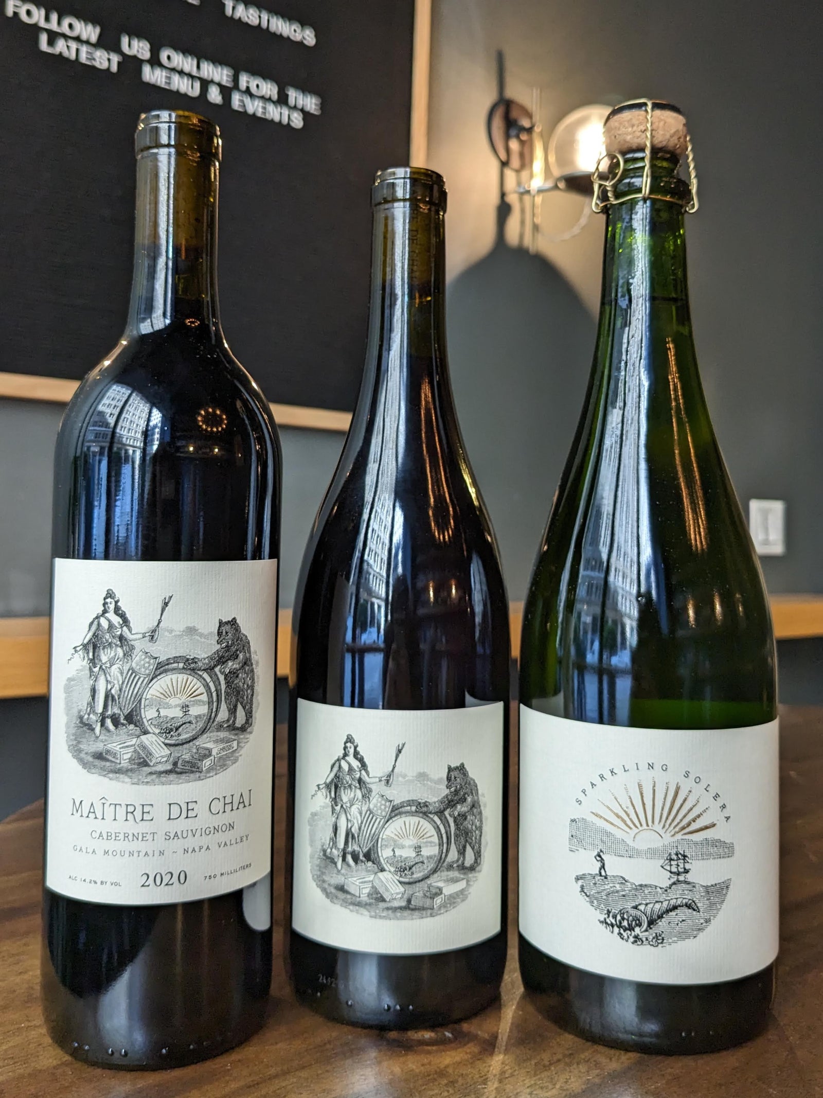 Three wine bottles displayed against a dark wall with signage: a dark wine bottle with white label on the left, a darker bottle in the center, and a g