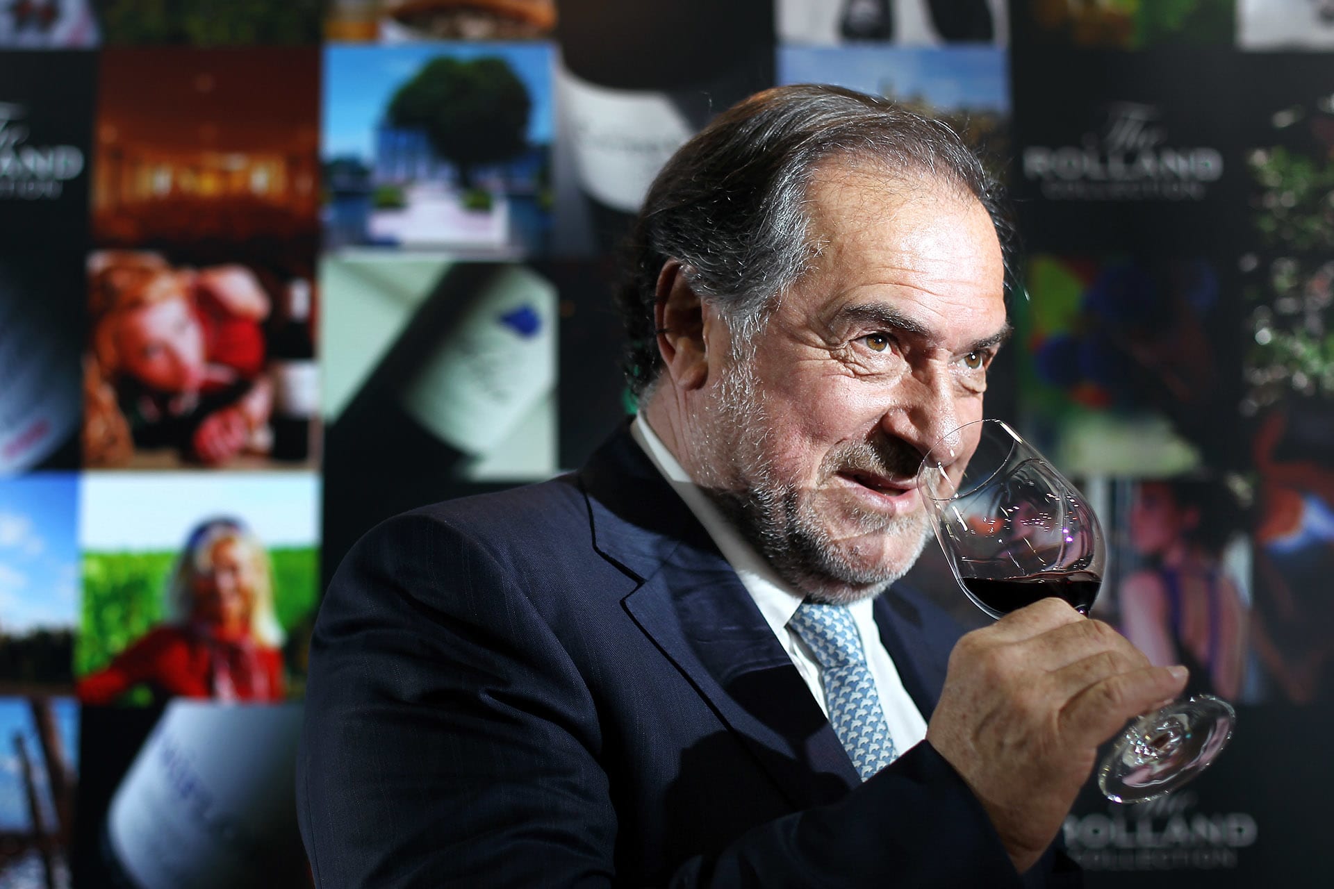 Man in dark suit and blue tie holding wine glass, with blurred wine images in background