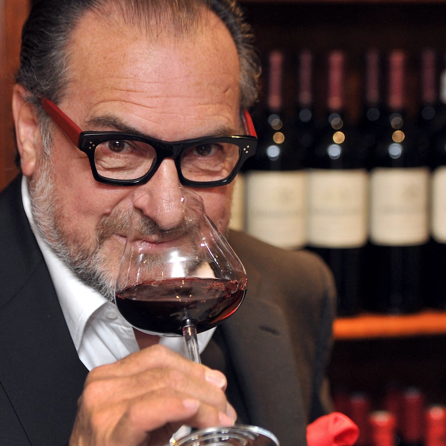 Man wearing black-framed glasses and holding a wine glass with red wine, tasting indoors
