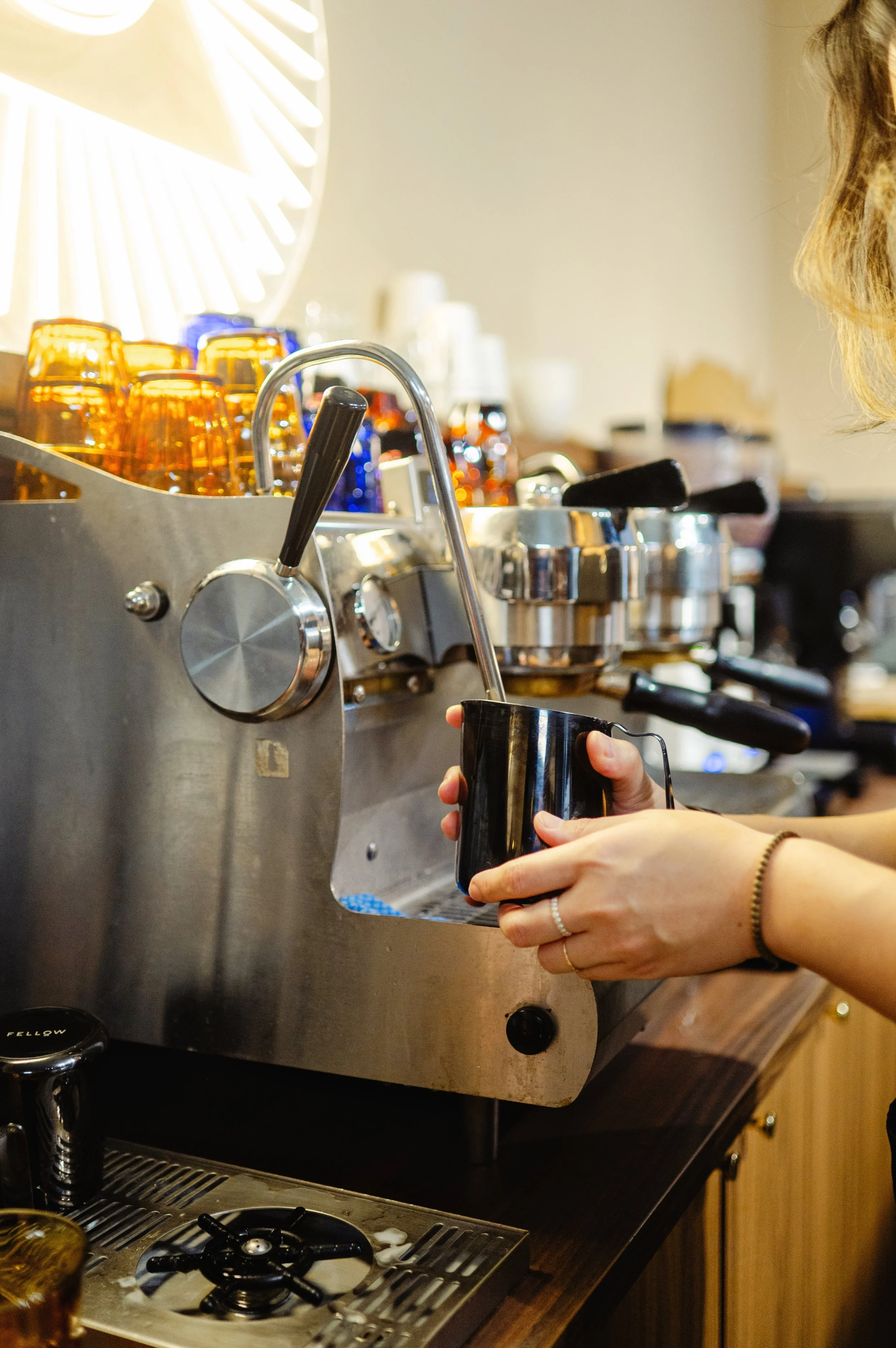 A barista steaming milk at a professional espresso bar with a stainless steel La Marzocco machine, grinders, and amber-colored bottles visible in the 