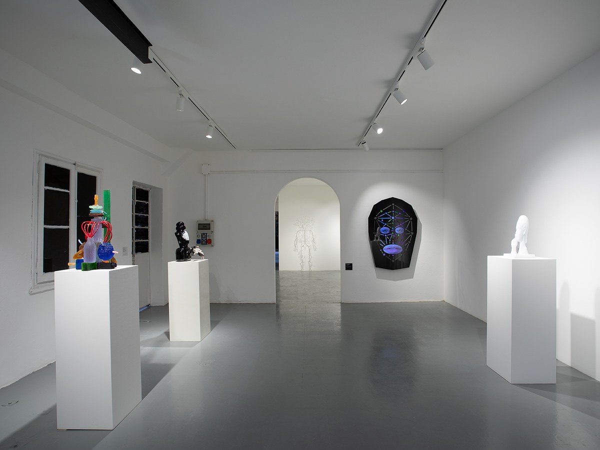 An interior view of a contemporary art gallery with white walls and grey floors, showcasing various glass sculptures on pedestals.