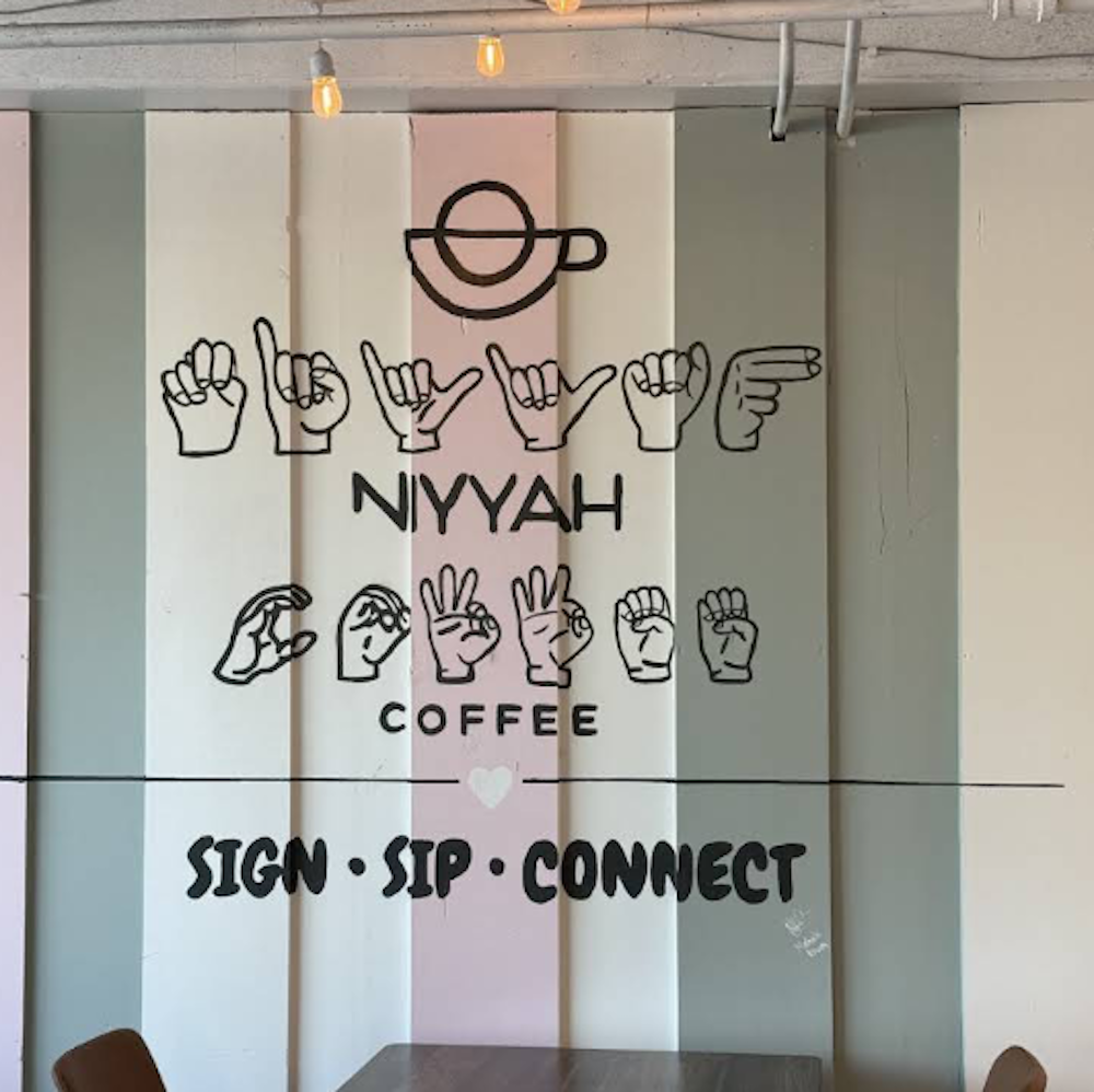 Interior wall signage for Niyyah Coffee showing the cafe name in both sign language hand symbols and text, with a coffee cup icon and tagline 'Sign • 
