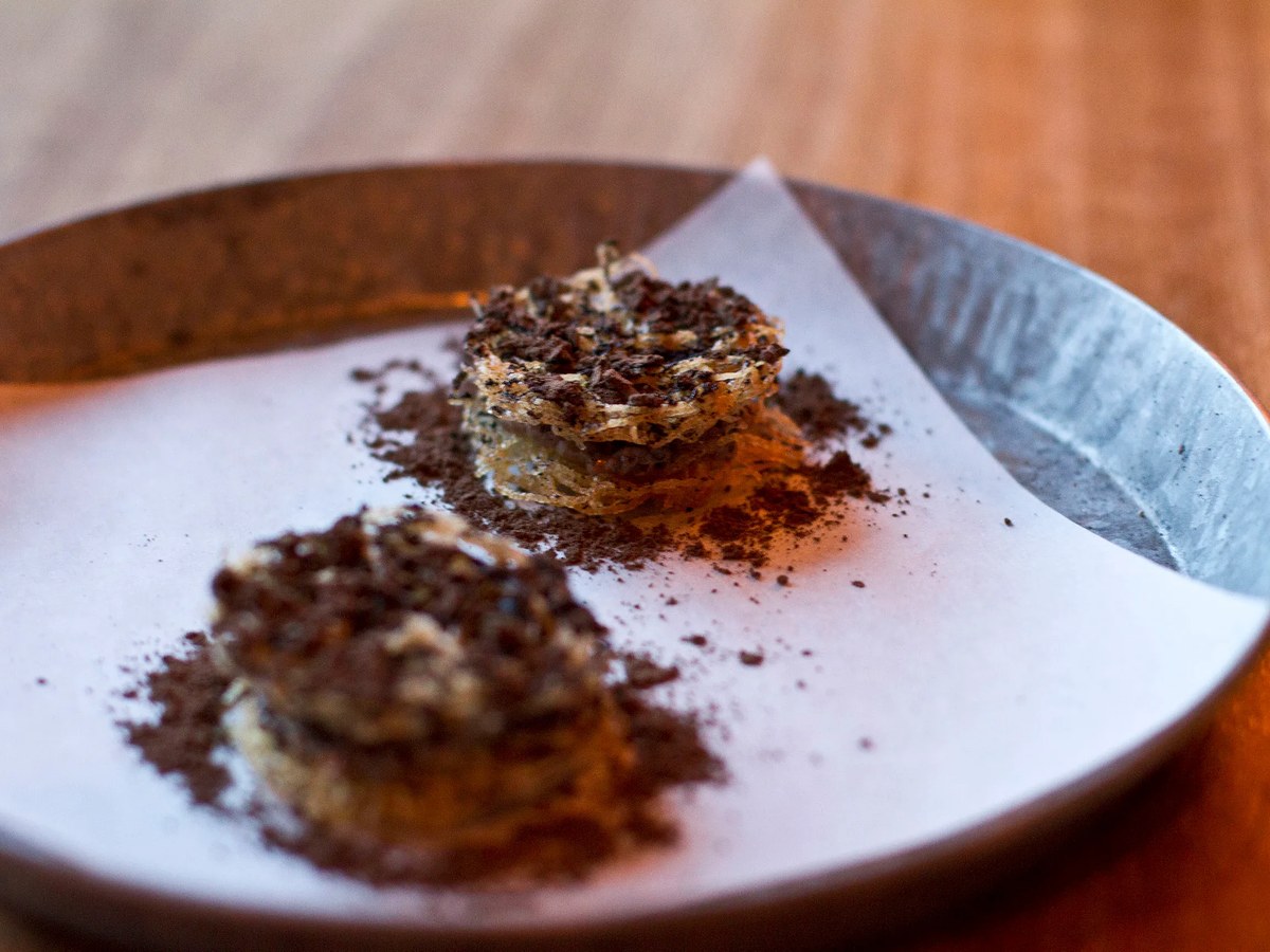 Two small fried potato haystack rounds, dusted with dark powder, are presented on a white paper on a ceramic plate.