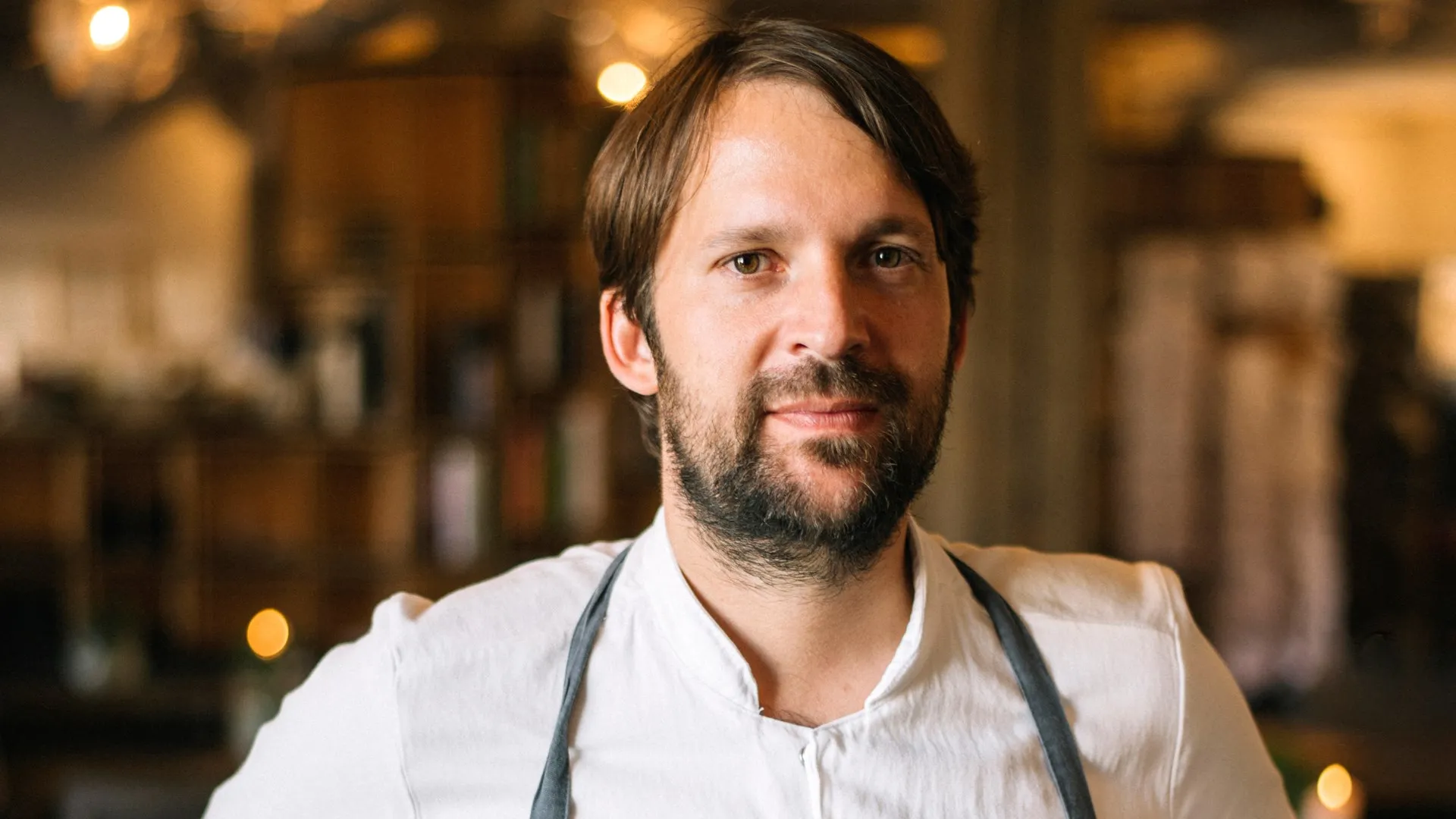 A man with a beard and dark hair, wearing a white shirt and a grey apron, looks directly at the camera.