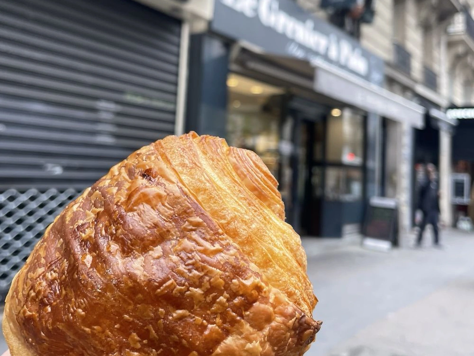 Close-up of a baked pie with crispy, textured pastry crust held up to a blurred urban street scene with storefronts in the background.