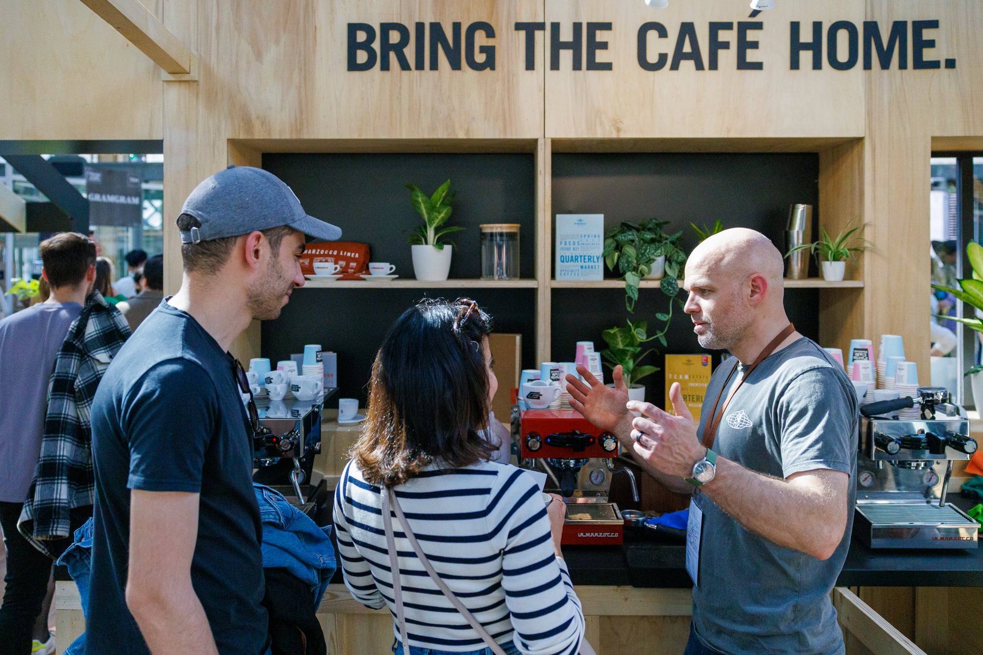A barista at a coffee trade show booth is demonstrating or explaining espresso equipment to two attendees in front of a display with shelving, plants,