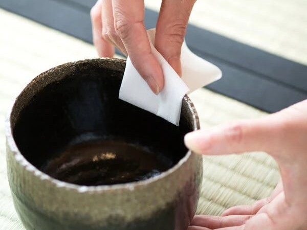 A close-up of hands performing a tea ceremony ritual, with one hand holding a white folded cloth (chakin) wiping the rim of a dark ceramic tea bowl