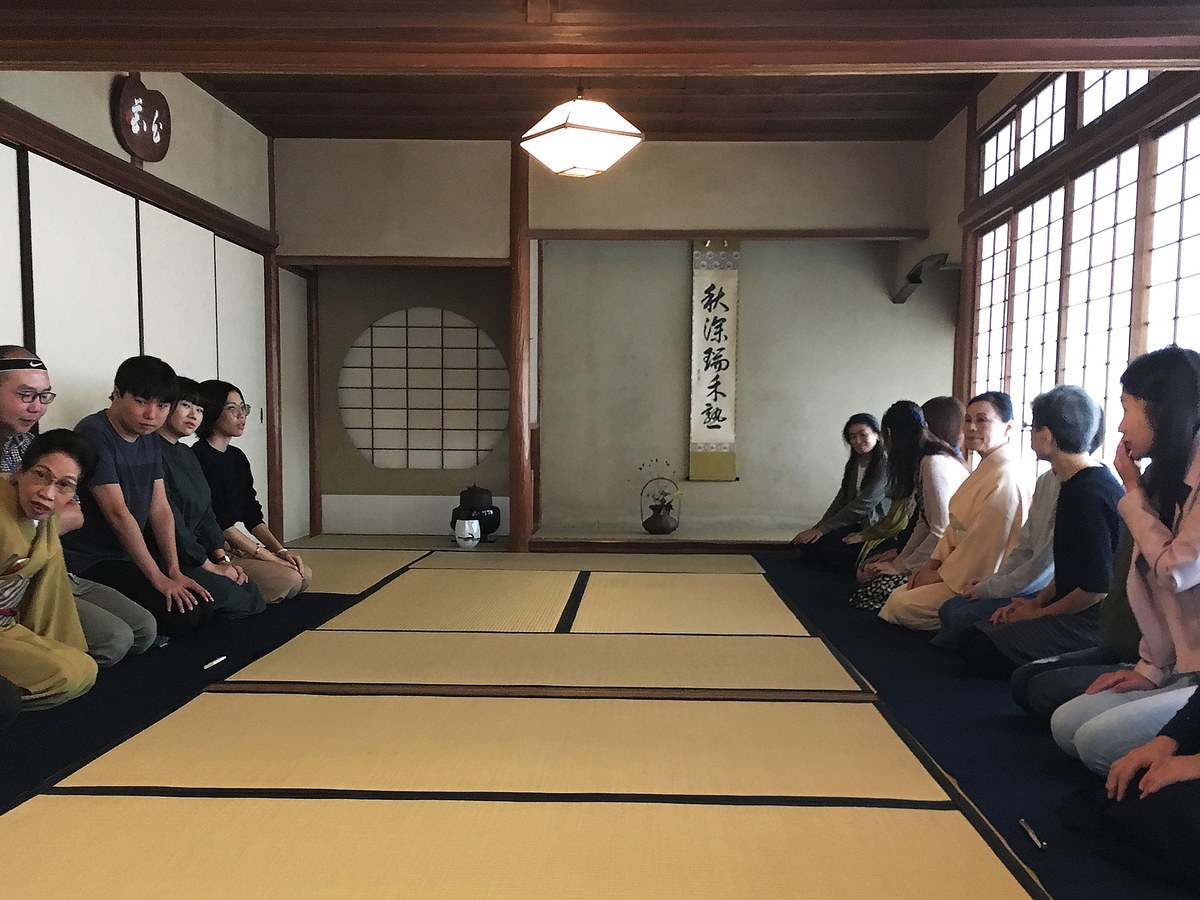 A group of participants sits in seiza on tatami mats in a traditional Japanese tea room, featuring shoji screens and a hanging scroll.