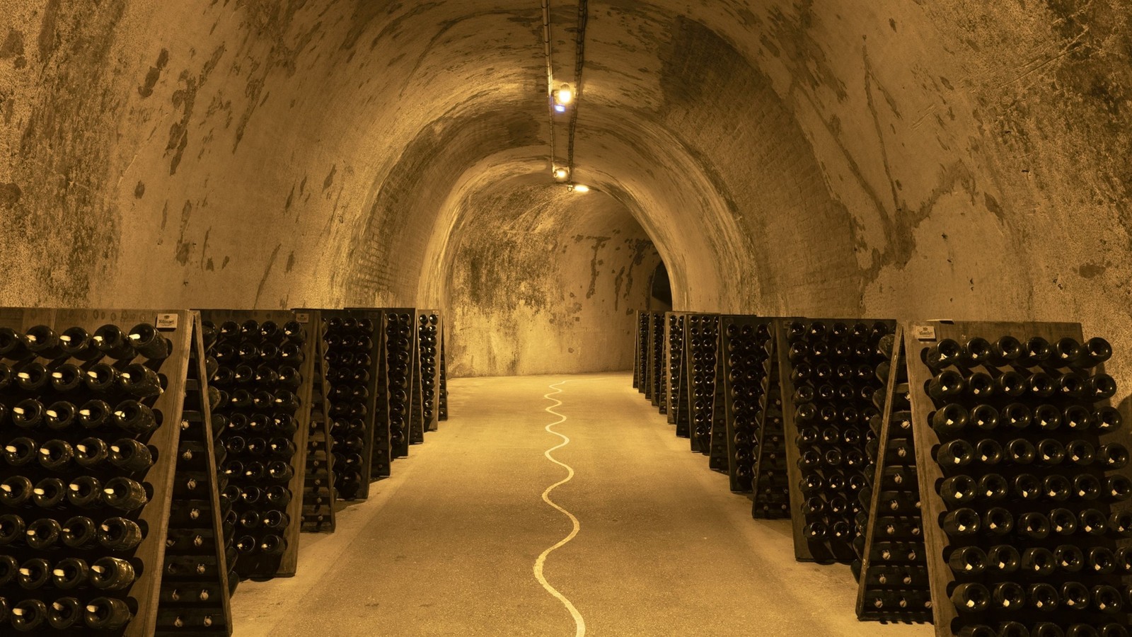 Interior view of a long arched underground tunnel lined with wooden racks holding dark wine bottles on both sides, with overhead lighting and a chalky stone ceiling.