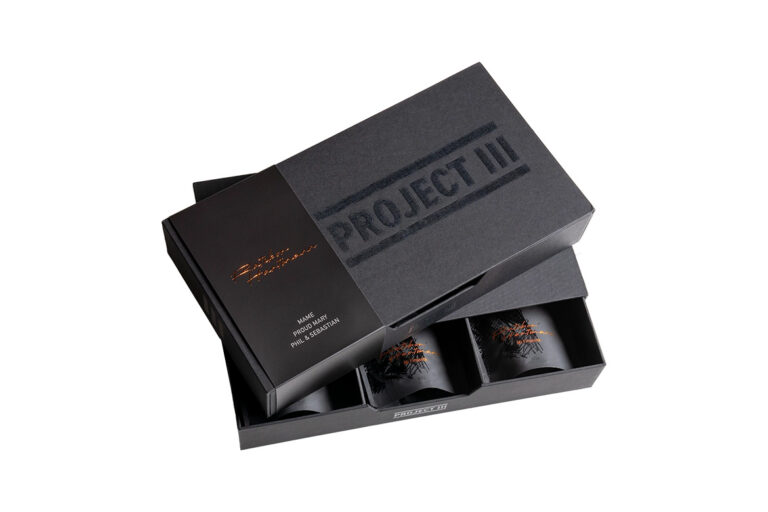 An open black box labeled Project III containing three black coffee containers with decorative designs and a black branded card with gold lettering.