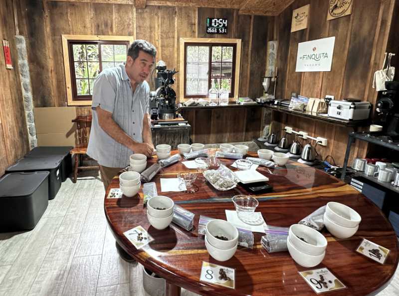 A man standing at a large round wooden cupping table with multiple white cups and coffee samples in a rustic wooden-walled lab