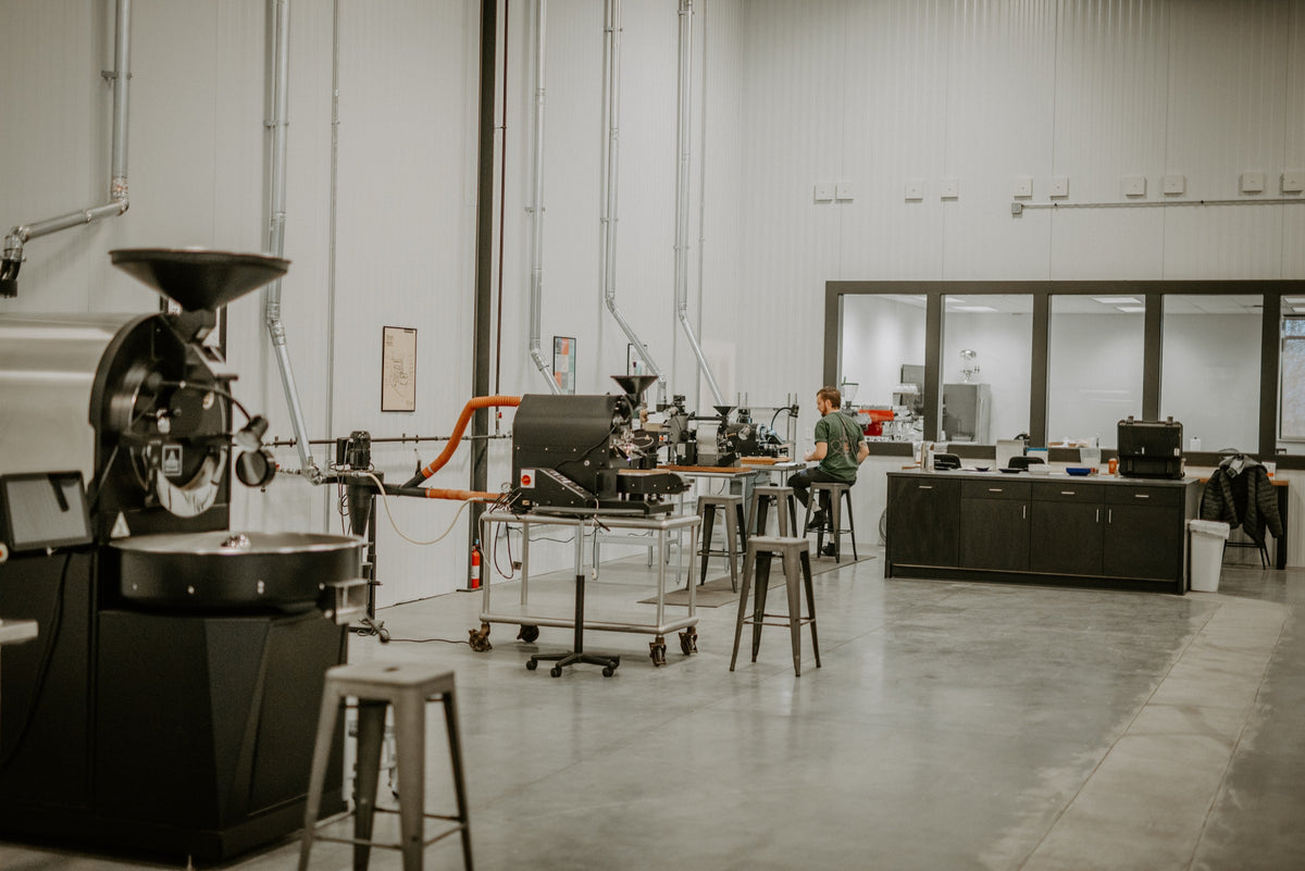 A coffee roasting facility with industrial roasting equipment on the left, a person working at a cupping/tasting station in the center, and an office 