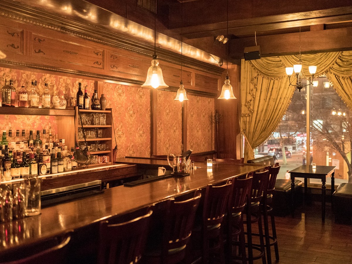 A dimly lit bar interior with a long wooden counter, bar stools, shelves of liquor bottles, and gold draped curtains next to a window.