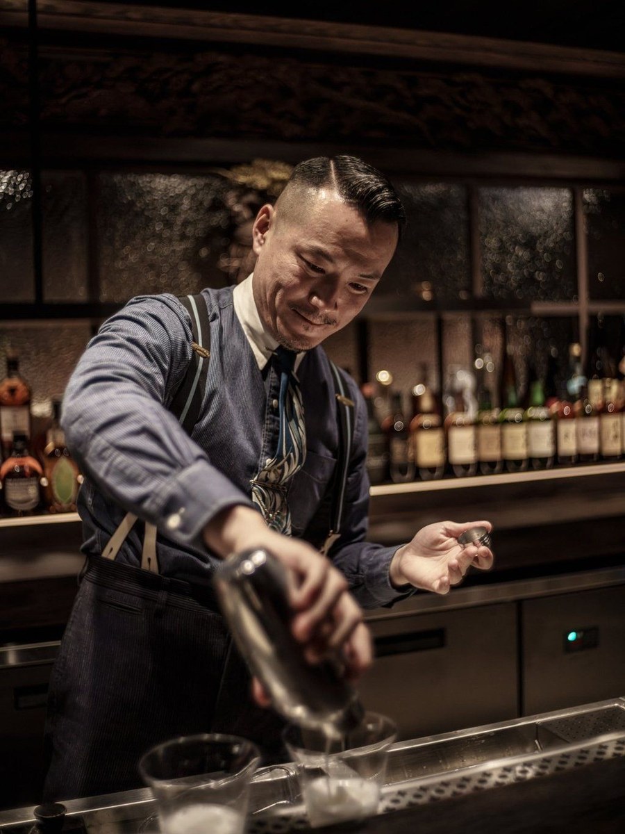 A male bartender in a blue pinstriped shirt and suspenders, with a neatly slicked-back haircut, pours liquid from a metal shaker into a glass behind