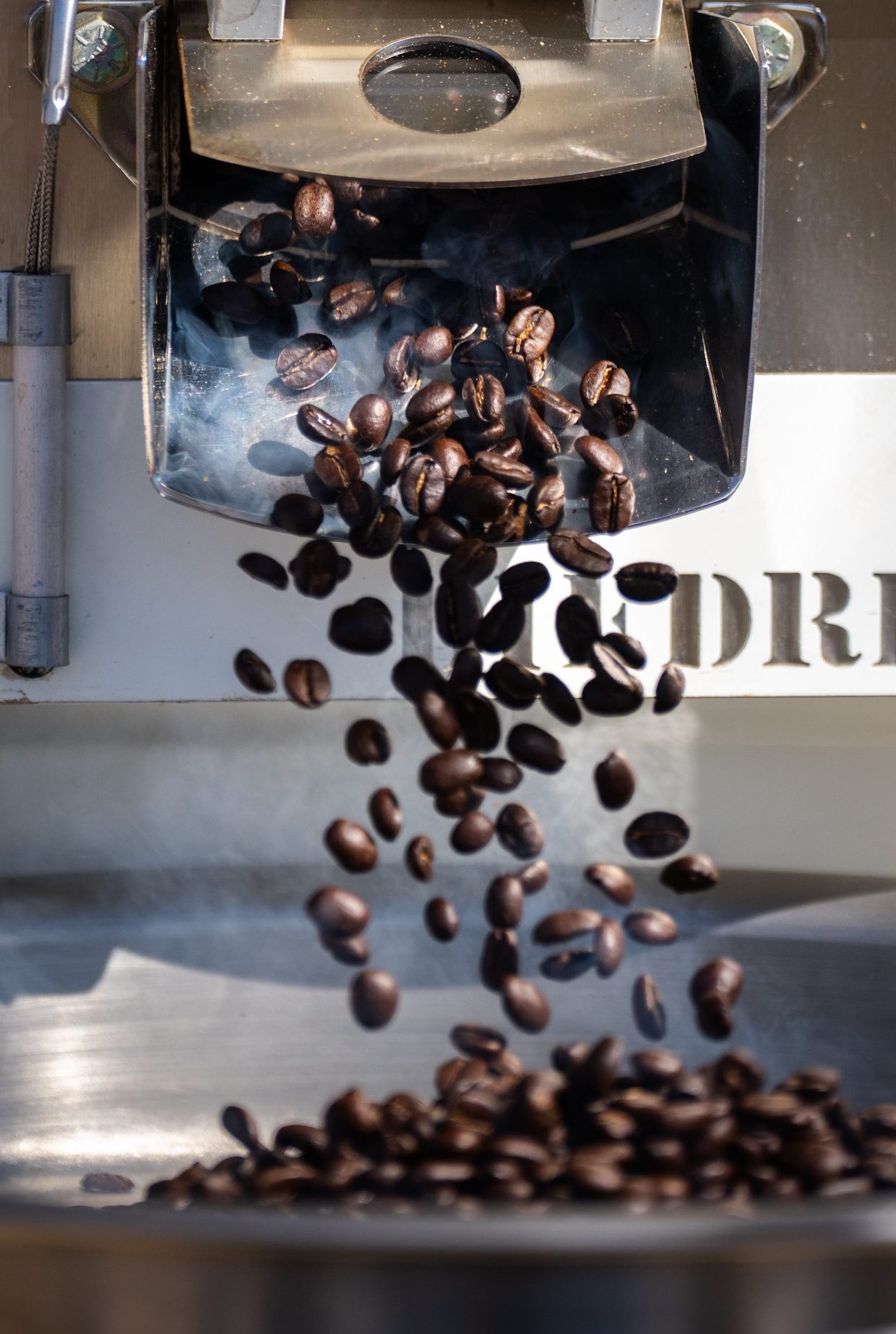 Coffee beans tumbling from a drum roaster during active roasting, with visible smoke/steam; the source page explicitly attests this is 'solar roasted 