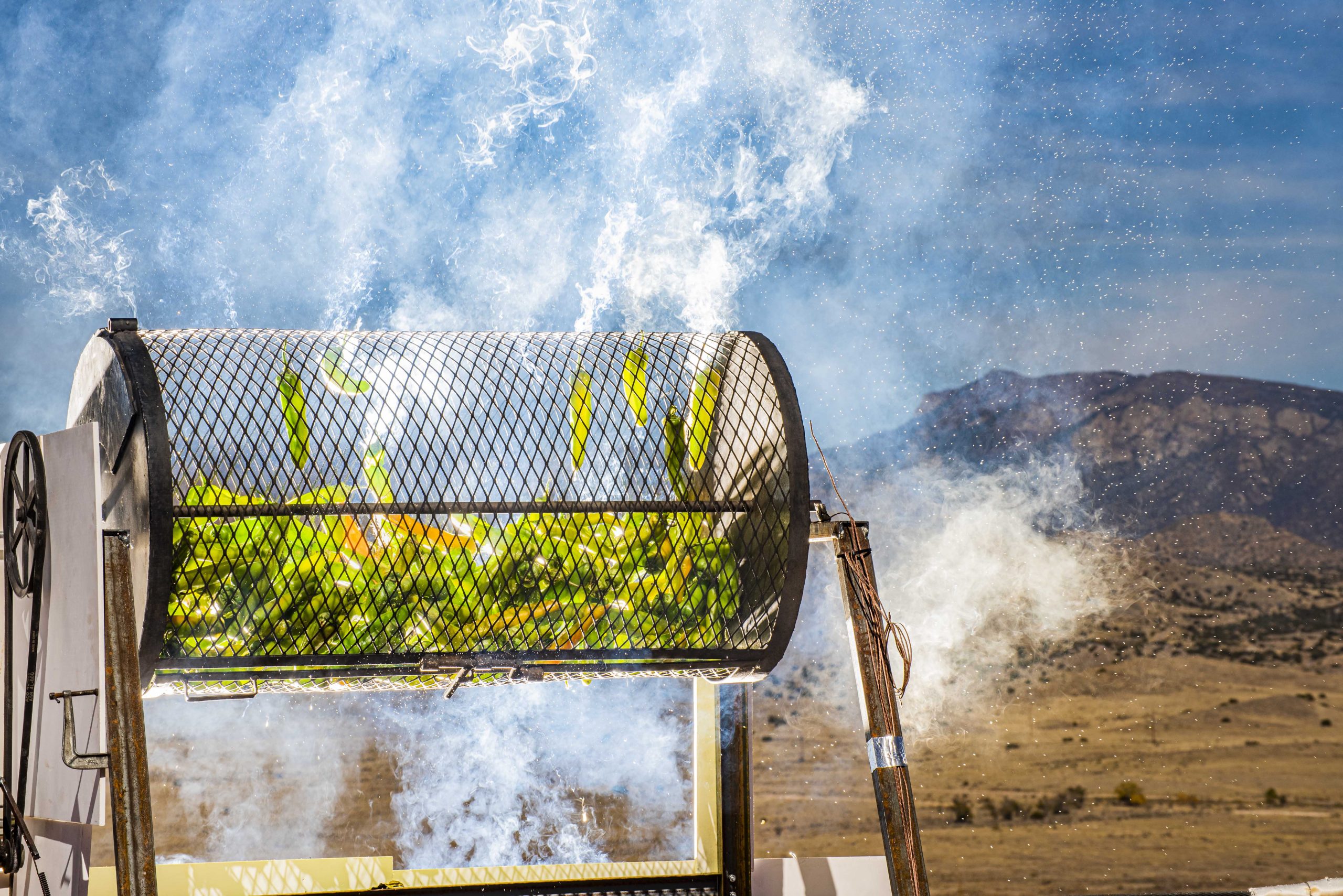 A rotating drum with yellow and green mesh containing coffee beans, set against a backdrop of steaming geothermal vents and barren volcanic terrain.