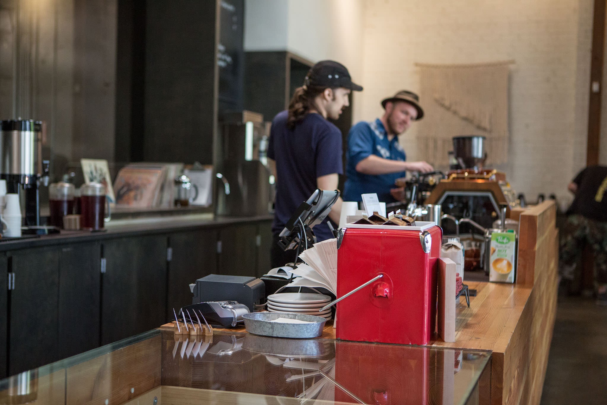 Coffee shop interior showing two baristas working behind a counter with espresso equipment, including what appears to be professional espresso machine