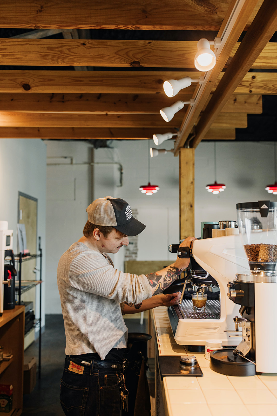 A barista in a baseball cap operates an espresso machine in a modern coffee shop with exposed wood beam ceiling and track lighting