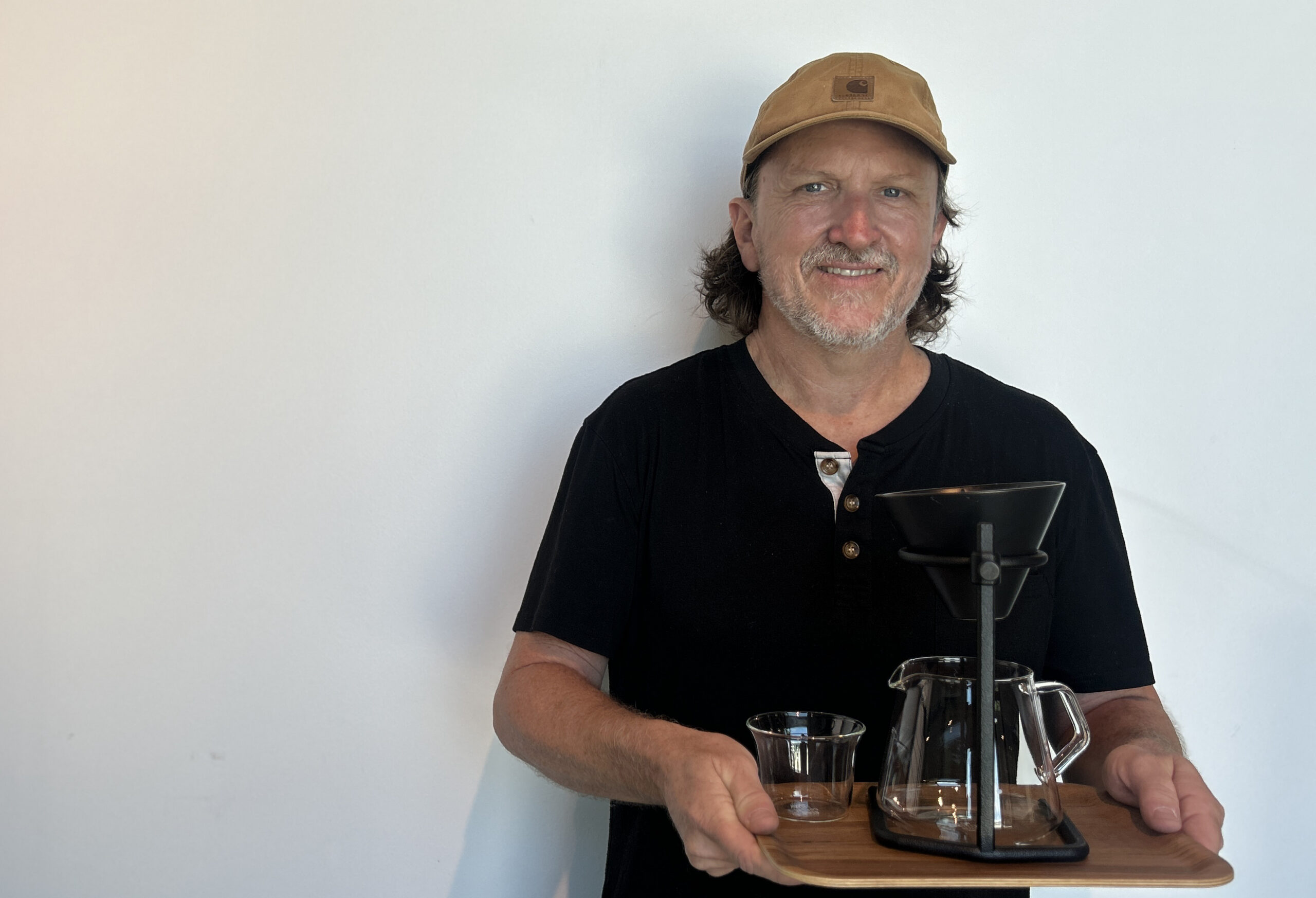 Portrait of a man in a tan cap and black henley shirt holding a wooden tray with coffee brewing equipment (carafe, pitcher, glass)