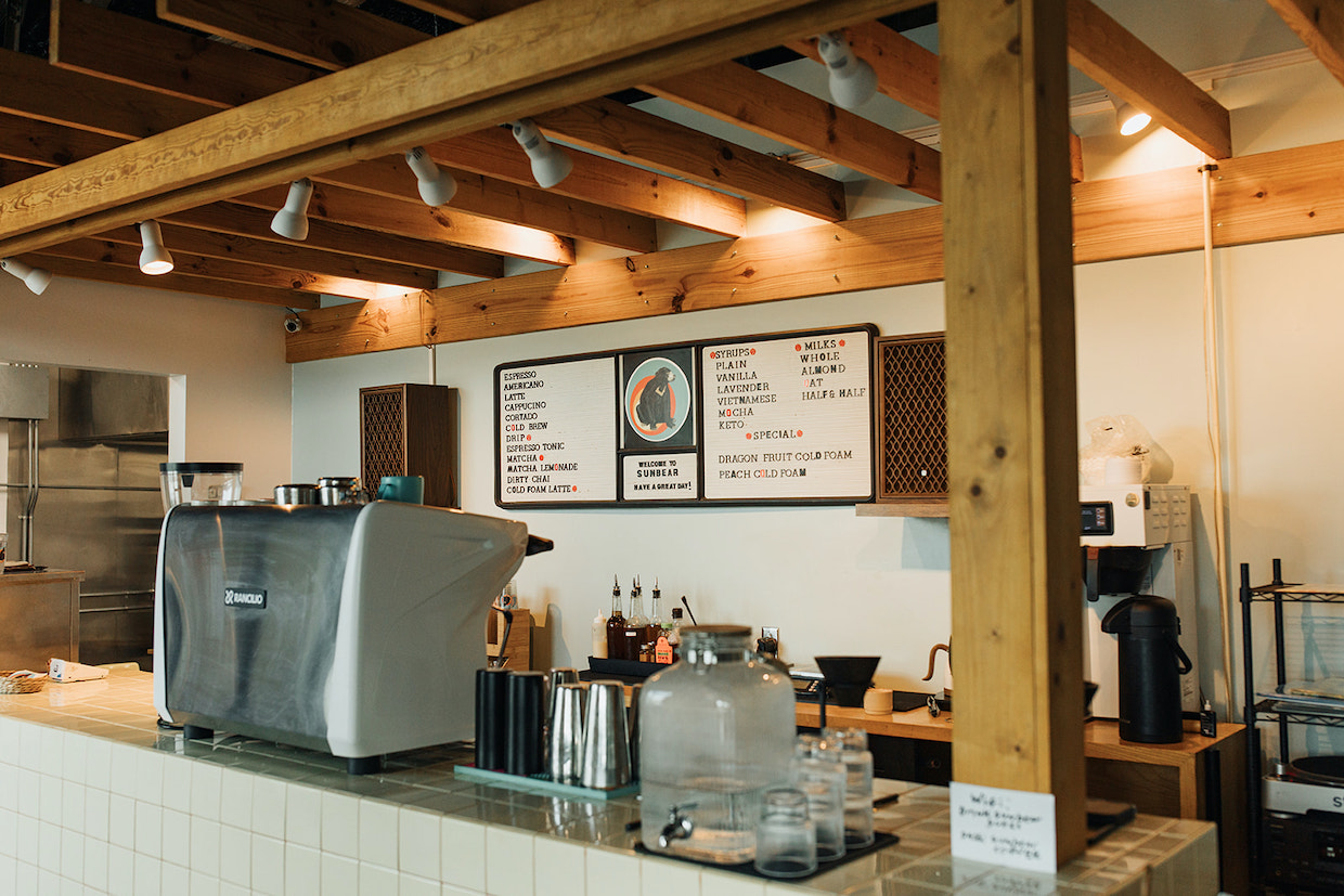 A modern coffee shop interior featuring an espresso bar with exposed wood beams, track lighting, menu boards, and professional equipment.