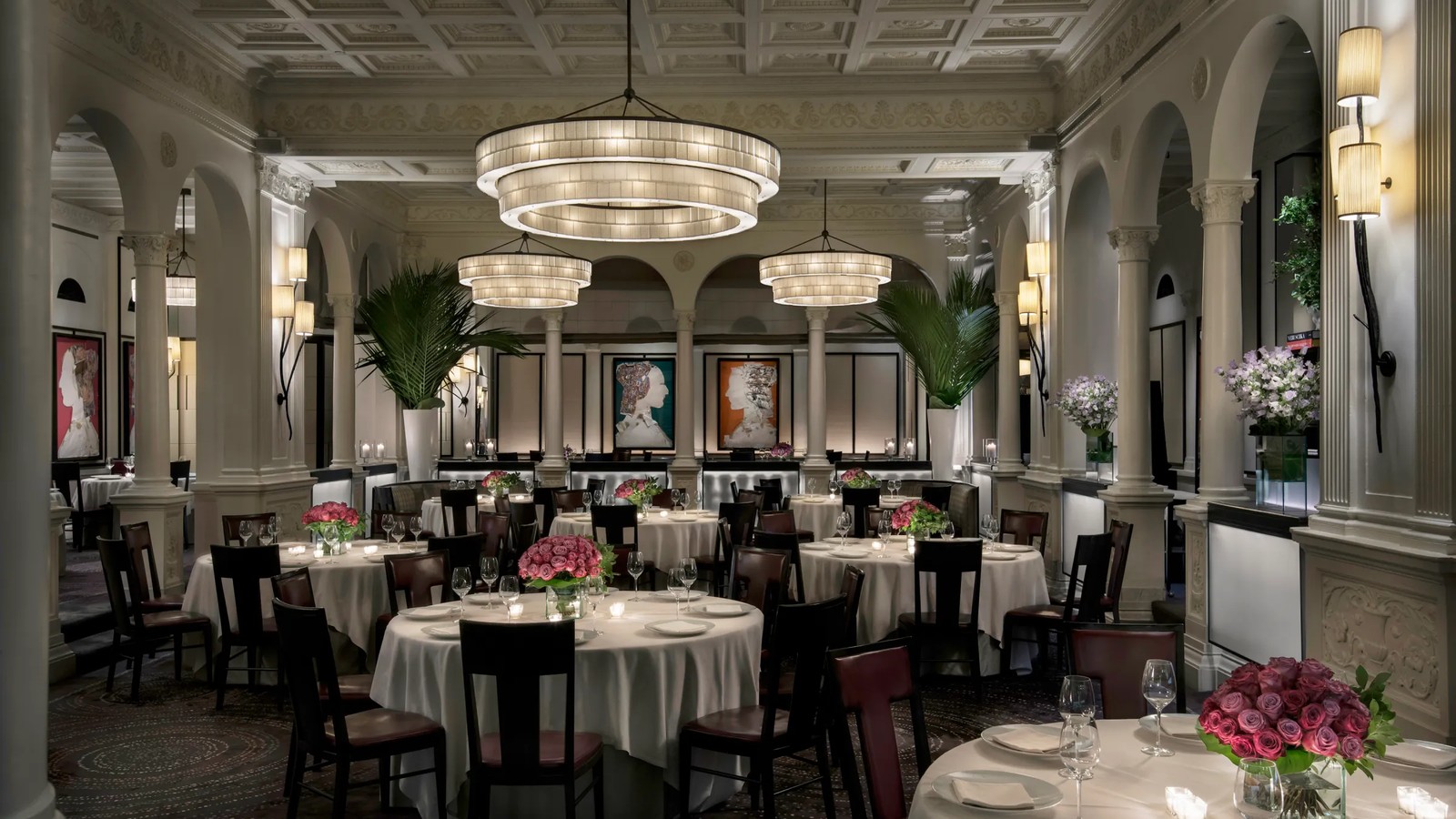 An elegant, high-ceilinged fine-dining room with white tablecloths, pink rose centerpieces, candlelight, tiered chandeliers, classical columns and arc
