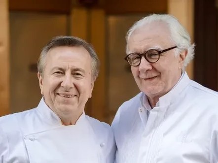 Two chefs in white jackets pose together outdoors.
