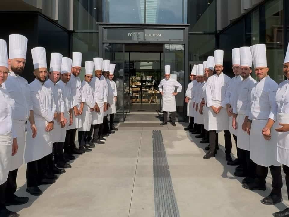 A large group of chefs wearing white coats and tall white chef hats lined up on both sides of a pathway outside a building entrance marked École Ducasse.