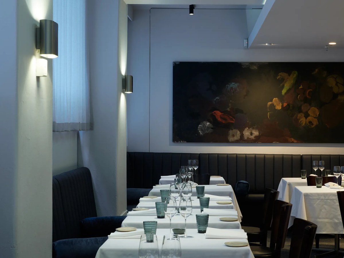 A contemporary fine-dining restaurant interior with white-linen tables, dark banquette seating, wine glasses, and a large dark floral painting on the