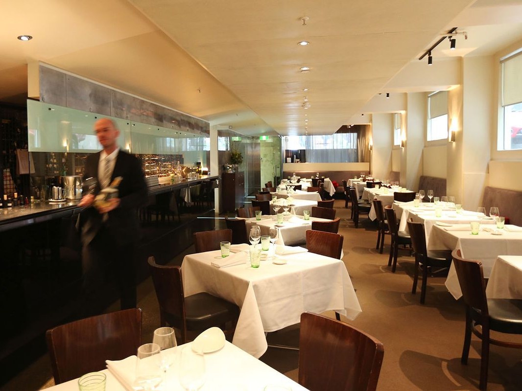 A fine-dining restaurant interior with white-tableclothed tables, dark timber chairs, warm lighting, a bar/wine area, and a motion-blurred suited