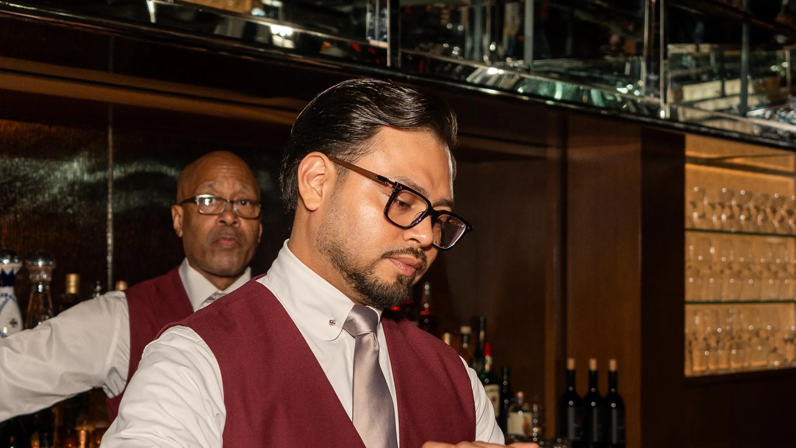 A bearded man wearing glasses, a white dress shirt, burgundy vest, and gray tie stands in a dimly lit restaurant with wine bottles and bar fixtures visible behind him.