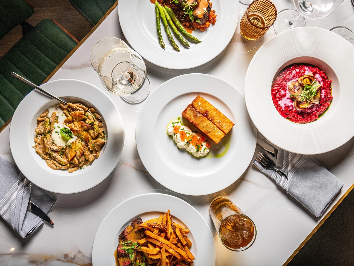 An overhead shot of a restaurant table set with multiple plated dishes including pasta, beet risotto, fried fish with roe, steak frites, and a meat