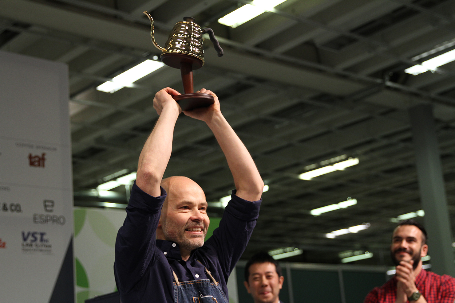 A man in a navy shirt holds a trophy aloft in an indoor venue with industrial ceiling and fluorescent lighting