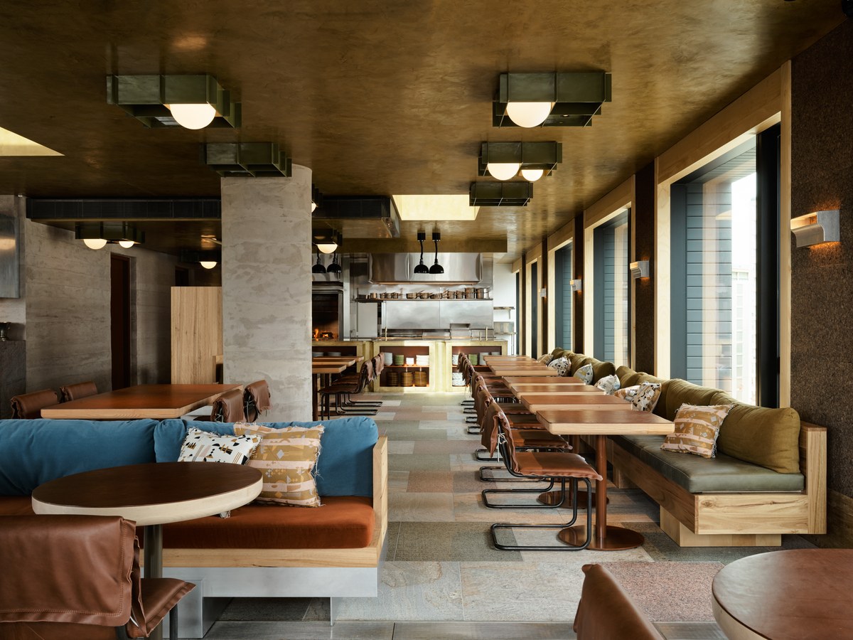 Modern restaurant interior with a gold-colored ceiling, concrete columns, booth seating, and an open kitchen.