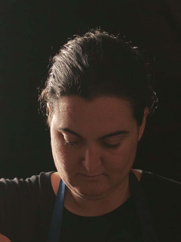 Portrait photograph of a person in chef's clothing, shown in profile with eyes downcast against a dark background.